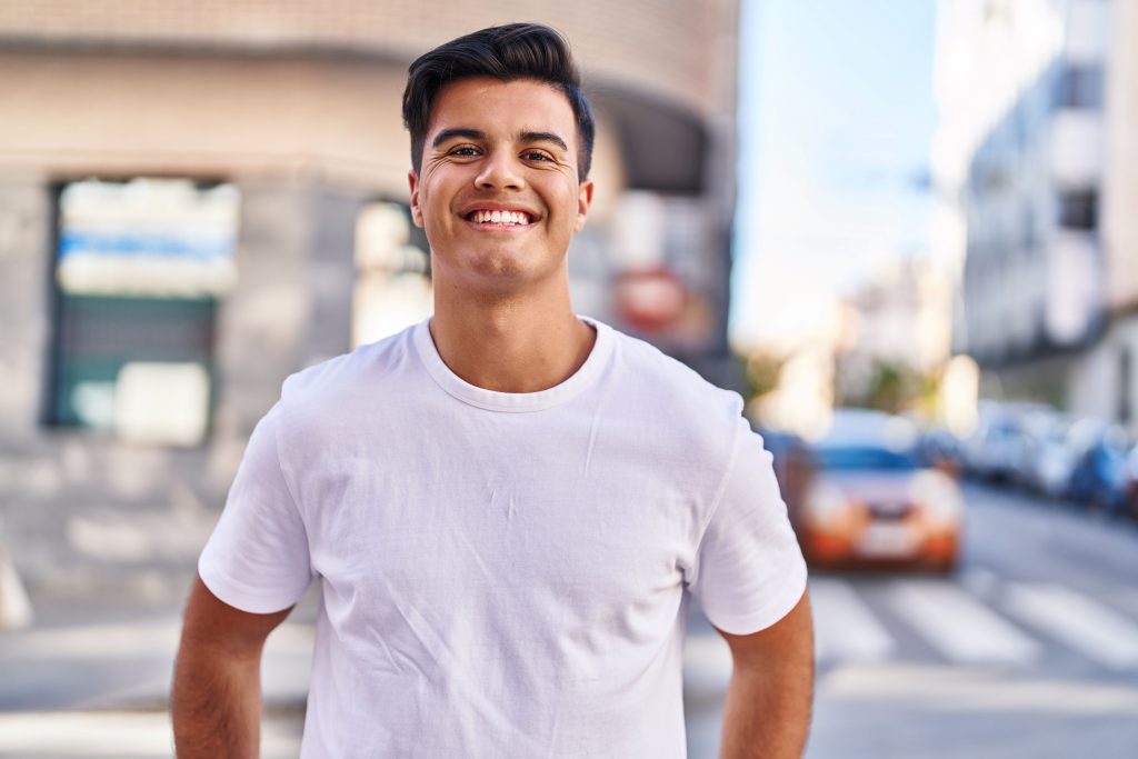 young man in white shirt smiles on sidewalk alongside a city street