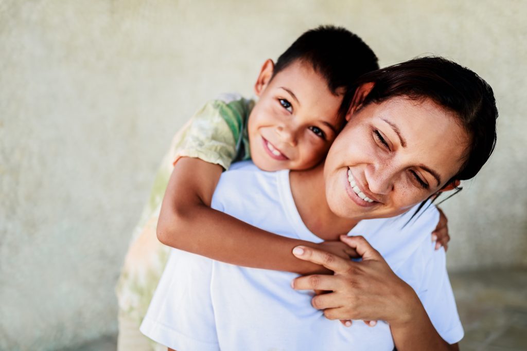 mother smiles as son hugs her from behind