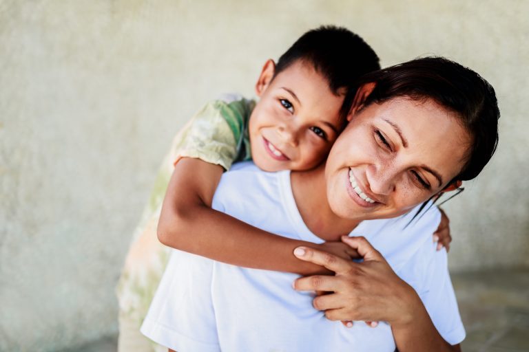 mother smiles as son hugs her from behind